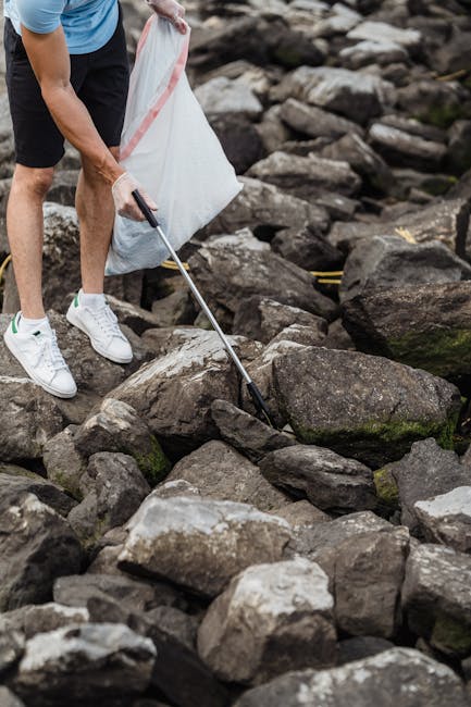 A person wearing white sneakers, black shorts, and a blue shirt is seen using a litter picker to collect small debris and trash from a rocky outdoor area. The individual is holding a large, partially filled white garbage bag, likely engaged in rubbish collection or environmental cleanup. The surrounding environment consists of numerous dark gray and brown rocks with irregular shapes, some with patches of green moss or lichen. The scene appears to be part of an outdoor natural setting, such as a riverside or rocky pathway, where waste might be collected for proper disposal through private rubbish removal services like Rubbish Removal St Johns Wood. The overall composition emphasizes hands-on waste collection, illustrating a manual approach to rubbish removal in an unmanaged outdoor area, relevant to sustainable waste handling methods and environmental responsibility.