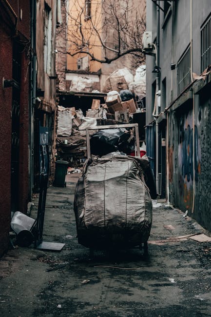 A narrow urban alleyway filled with various waste materials, with a large, black wheeled refuse sack placed centrally in the foreground. Behind the sack, there are piled cardboard boxes, plastic bags, and miscellaneous rubbish stacked against a weathered wall at the back of the alley. The left side features a brick wall with graffiti and a small black trash bin on the ground, while the right side shows a grey building with barred windows and an electrical box mounted on the wall. The ground is paved with uneven asphalt, stained and scattered with debris, indicative of an area used for informal waste disposal. Bare tree branches extend overhead, and the overall lighting is subdued with natural daylight filtering through the confined space, suggesting an external but enclosed environment. This scene exemplifies an area where independent waste collection and rubbish removal services like those provided by Rubbish Removal St Johns Wood are often utilized for managing refuse outside of regular municipal collection routes, highlighting the need for professional clearance in such cluttered urban settings.