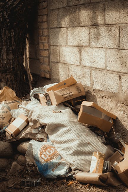 A person wearing white sneakers, black shorts, and a blue shirt is seen using a litter picker to collect small debris and trash from a rocky outdoor area. The individual is holding a large, partially filled white garbage bag, likely engaged in rubbish collection or environmental cleanup. The surrounding environment consists of numerous dark gray and brown rocks with irregular shapes, some with patches of green moss or lichen. The scene appears to be part of an outdoor natural setting, such as a riverside or rocky pathway, where waste might be collected for proper disposal through private rubbish removal services like Rubbish Removal St Johns Wood. The overall composition emphasizes hands-on waste collection, illustrating a manual approach to rubbish removal in an unmanaged outdoor area, relevant to sustainable waste handling methods and environmental responsibility.