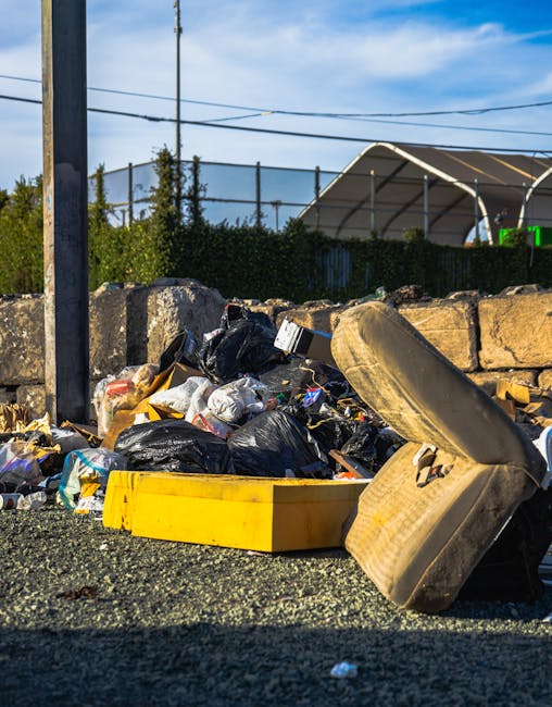 A container of mixed waste is overflowing onto a gravel surface, with various black and clear plastic rubbish bags, cardboard, and discarded items spilling out. Prominent in the foreground is an old, dirty car seat with beige upholstery, partly tilted and resting against the pile of refuse. Behind the rubbish, there is a low stone wall made of rough, uneven stones, and beyond that, a hedge and some trees are visible. To the left, a metal utility pole with power lines extends upwards, crossing the bright blue sky with some scattered clouds. The scene appears to be outdoors on an everyday street or roadside area, where waste has been temporarily dumped. The background includes a sports or event facility with a curved, white canopy structure and a partially visible green fence, suggesting the location might be near a public or recreational area. The natural daylight illuminates the scene, highlighting the textures and colors of the trash and surroundings, subtly indicating the use of private or independent rubbish disposal services such as those offered by Rubbish Removal St Johns Wood for local waste collection needs.
