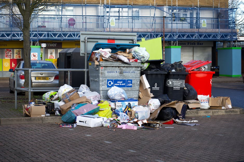 A collection of overflowing rubbish bins and scattered waste sitting on a paved urban street corner. The scene includes multiple black, grey, and red bins, some with lids open, revealing materials such as cardboard boxes, plastic bags, paper, and miscellaneous packaging. The waste piles extend across the sidewalk and onto the curb, with various discarded items like flattened boxes, plastic containers, and crumpled paper visibly spilling onto the pavement. In the background, there are storefronts with signage, a parked car to the left, and a metal railing separating the waste area from the street. Behind the waste, a building under renovation is visible, covered with blue scaffolding and netting. The environment appears to be during daylight, with overcast weather suggesting natural, diffuse lighting. This scene illustrates an example of excess waste typical in private collection or on-site rubbish clearance scenarios managed by companies such as Rubbish Removal St Johns Wood, highlighting the importance of timely rubbish removal services in urban areas.
