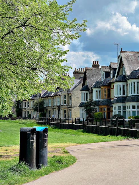 A row of traditional Victorian-style terraced houses with brick facades and bay windows, situated behind a black metal fence, are visible in the background under a partly cloudy sky. In the foreground, a grassy area with a wide pathway runs alongside the houses, with two black waste bins placed on the edge of the grass near a large, leafy green tree whose branches extend across the upper left corner of the image. The scene appears to be in a residential neighborhood, illustrating an outdoor setting suitable for private waste collection or rubbish removal services. The overall atmosphere is bright and clear, emphasizing the outdoor environment and the potential for rubbish clearance in such areas, supported by Rubbish Removal St Johns Wood's expertise in managing waste outside of traditional municipal collection methods.