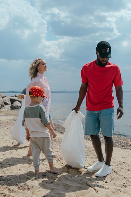 A group of three people, including an adult male and two children, are participating in a beach clean-up activity during daytime. The man, positioned to the right, is wearing a black cap, a red t-shirt, and light blue denim shorts, and is holding a white rubbish bag while standing barefoot on the sandy shoreline. To his left, a young boy in a grey t-shirt, beige pants, and a red cap with white polka dots is also holding a white rubbish bag, with his back partially turned. Behind them, a young woman with curly blonde hair, dressed in a light pink shirt, is observing the scene with her face turned slightly to the left. The background features a calm body of water extending to the horizon, with large rocks along the shoreline and a partly cloudy sky overhead. The environment suggests a voluntary effort to remove waste from the beach, indicative of private or community-based rubbish collection services, such as those offered by Rubbish Removal St Johns Wood, supporting alternative and environmentally conscious waste handling.