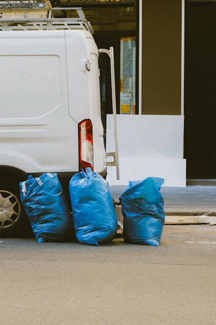 A white commercial van parked on a city street with its rear door closed, positioned next to a sidewalk. In front of the van, three large blue rubbish bags made of thick, durable plastic are stacked on the paved surface, filled with waste material. The bags are slightly crumpled and appear to be sealed, ready for collection. Behind the van, there is a modern building with large glass windows and a black-framed entrance, along with a white partition or barrier near the doorway. The environment suggests a typical urban setting, possibly outside a commercial or residential property, with natural daylight illuminating the scene. The presence of the bags indicates an instance of private waste disposal, consistent with independent rubbish removal services such as those offered by Rubbish Removal St Johns Wood. This setup exemplifies a common method of on-site waste clearance prior to scheduled collection or alternative rubbish handling. The scene overall highlights an accessible, straightforward approach to rubbish disposal outside a typical urban premises.